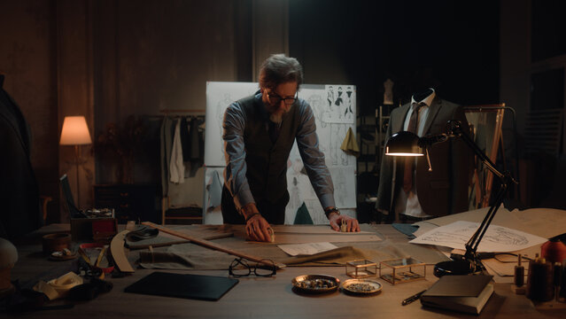 Male mature tailor in luxury designer atelier or tailoring studio. He works with sketch of future garment on table with fabric and tools. Mannequin with tailored suit. Fashion and hand craft concept.