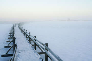 A snowy wooden boardwalk on a frozen lake to a birdwatching tower in the evening sun and mist