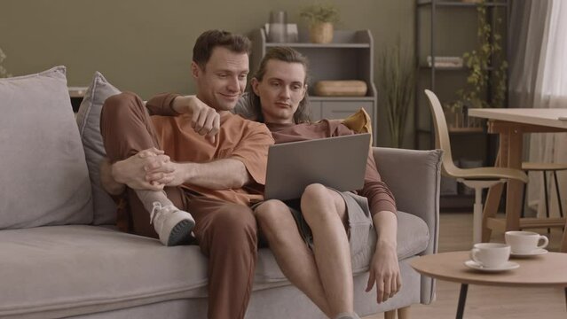 Romantic gay couple sitting on comfortable couch in living room while watching movie on laptop together