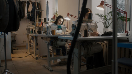 Female seamstresses sitting at sewing machines and working on custom apparel. Measure tapes and laptop with sketch of wedding suit at the table. Atelier or sewing workshop. Concept of fashion.