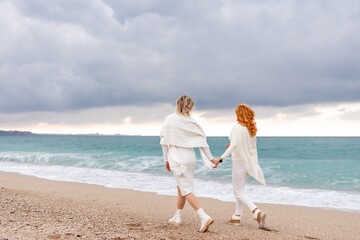 Fototapeta na wymiar Women sea walk friendship spring. Two girlfriends, redhead and blonde, middle aged walk along the sandy beach of the sea, dressed in white clothes. Against the backdrop of a cloudy sky and the winter 