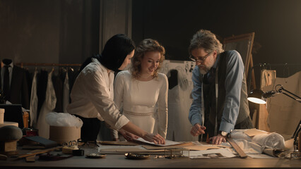 Cinematic shot of tailors and woman discussing sketches of wedding dress. Bride on fitting wedding dress in luxury designer atelier or tailoring studio. Concept of fashion, handmade and couturier.