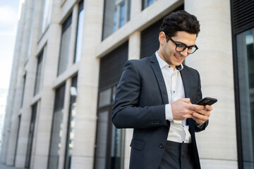 A young businessman in a formal suit on the way to the office writes a message to a colleague on the Internet using a smartphone. Education, business, the concept of an office employee.