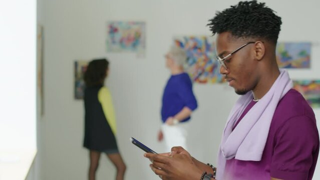 Waist up shot of African American man taking picture of artwork with smartphone while visiting exhibition in gallery