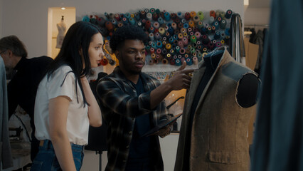 African American fashion designer talks with seamstress in atelier and shows sketch of future suit on digital tablet computer. Mannequin in unfinished tailored suit. Concept of fashion and hand craft.