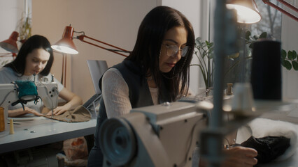 Female tailor in glasses sits at sewing machine and works on custom apparel. Seamstress colleagues working by the table with laptop at background. Atelier or sewing workshop. Concept of fashion.