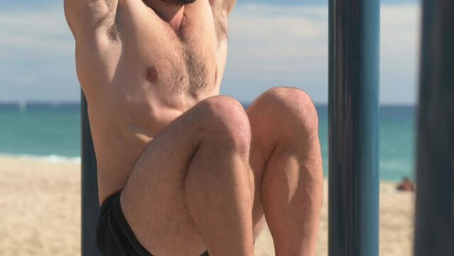 Shirtless young man doing sit-ups on a bar in a park at the beach.