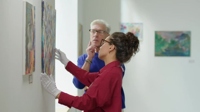 Female gallery curator in gloves hanging painting on wall and discussing it with mature male colleague while working together on exhibition