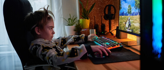 Image of immersed teenage gamer boy playing video games on computer in dark room wearing headphones and using backlit colorful keyboard © stenkovlad