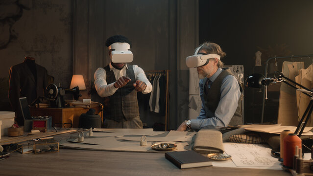 Multi ethnic tailors wearing VR headset with wireless controllers. They working in luxury atelier or tailoring studio and creating business suit in virtual reality. Concept of technologies in business