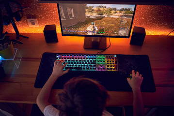 Image of immersed teenage gamer boy playing video games on computer in dark room wearing headphones and using backlit colorful keyboard