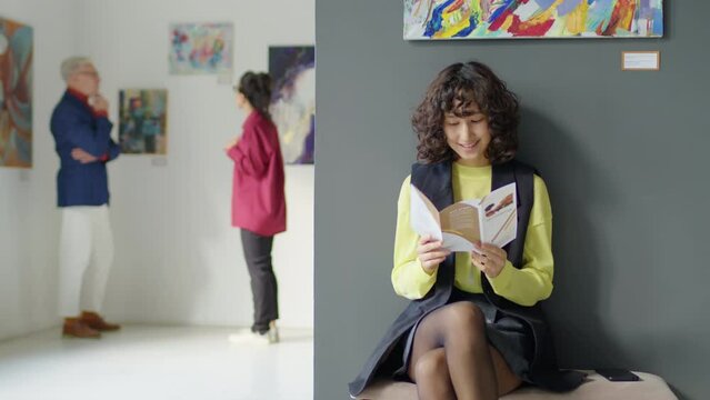 Tracking shot of young woman sitting on bench in exhibition hall of art gallery and reading booklet
