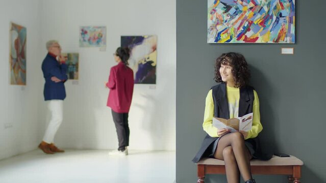 Tracking shot of young female visitor sitting on bench in art gallery and reading information about exhibition in booklet