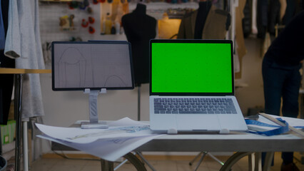 Dolly shot of laptop with green screen and digital tablet computer with sketch on table in atelier workshop. Tailor and seamstress work on background. Concept of fashion and technologies in business.
