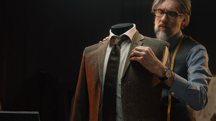 Male mature tailor works on elegant business or wedding suit. Mannequin with tailored shirt, tie and jacket in stylish luxury designer atelier or tailoring dim studio. Fashion and hand craft concept.