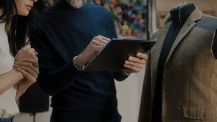 Male fashion designer shows sketch of future suit jacket to seamstress on tablet computer and discusses tailoring process. Mannequin in atelier workshop. Fashion and hand craft concept. Close-up shot.
