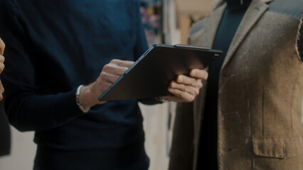 Male fashion designer shows sketch of future suit jacket to seamstress on tablet computer and discusses tailoring process. Mannequin in atelier workshop. Fashion and hand craft concept. Close-up shot.