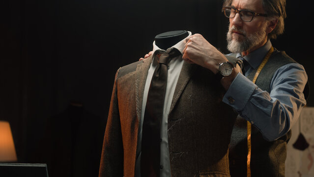 Male mature tailor works on elegant business or wedding suit. Mannequin with tailored shirt, tie and jacket in stylish luxury designer atelier or tailoring dim studio. Fashion and hand craft concept.