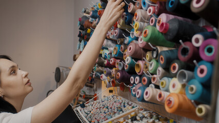 Female fashion designer chooses color of threads for tailoring new elegant wedding suit. Woman takes skeins of threads while working on custom apparel. Atelier or sewing workshop. Concept of fashion.