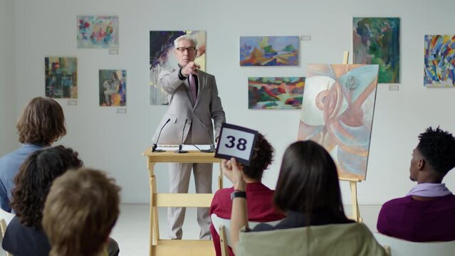 Male auctioneer with gray hair standing by rostrum on stage and calling out bids while selling artwork on auction