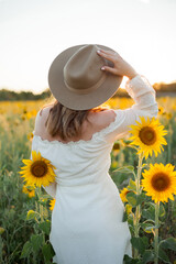 Portrait of beautiful young woman 33 years old in hat in sunflower field at sunset. Happy model in...