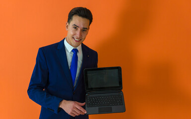 waist up photo of a dapper man dressed in suit holding a laptop while looking at camera ahead an orange background