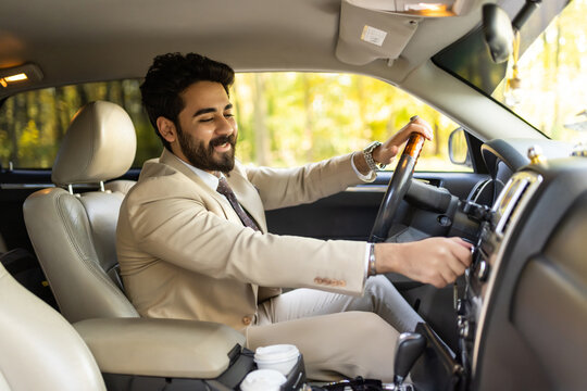 Happy arab young man businessman sitting inside luxury automobile, turning on music on dashboard before driving, handsome arabic man having car trip, side view, copy space