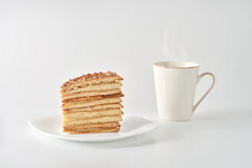 A piece of cake from a variety of cakes with honey in the composition on a white plate on a white background. Next to it is a cup with a hot drink. Steam rises from the drink.