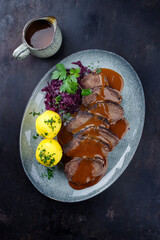 Traditionell geschmorter Sauerbraten vom Rind mit Blaukraut und Kartoffelklößen in würziger Dunkelbiersoße serviert als Draufsicht auf einem Nordic design Teller
