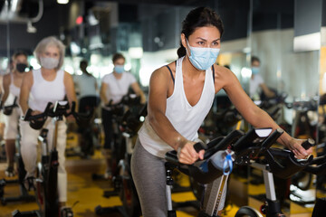 Portrait of young adult woman wearing face mask for disease protection training on stationary bike workout in gym
