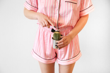 a girl in pink pajamas holds a cactus and shaves its thorns. Body hair problem, stubble after shaving. Laser hair removal, waxing, shugaring, electrolysis. Shaving with a machine, depilation of the bi - obrazy, fototapety, plakaty