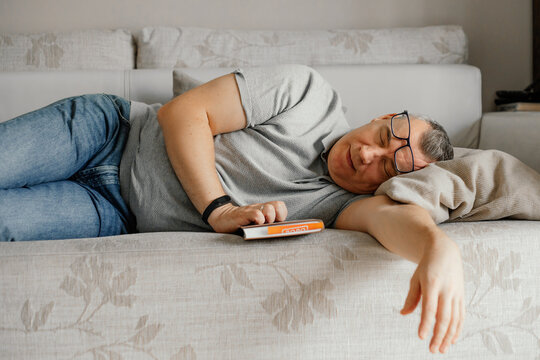 Aged Man With Glasses Fall Asleep Reading Book On Comfortable Couch In Home Room. Daytime Sleep, Napping.