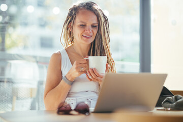 Attractive smiling woman with dreadlocks enjoying coffee while working in cafe.