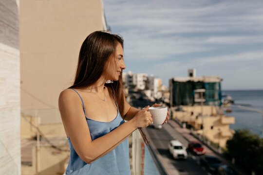 Outdoor photo of romantic pretty woman with dark long hair wearing blue shirt enjoying sunny morning on the balcony with coffee and looks forward.
