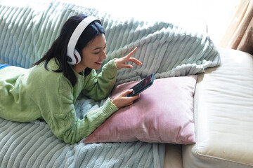 Image of happy biracial woman in headphones using smartphone lying on sofa at home, copy space - Powered by Adobe