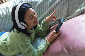 Image of happy biracial woman in headphones using smartphone relaxing on sofa at home, copy space - Powered by Adobe