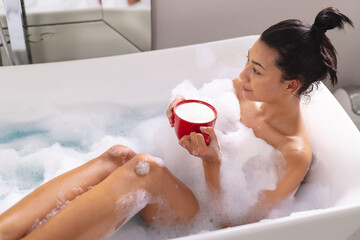 Image of biracial woman relaxing with cup of tea in bathtub in bubble bath in bathroom