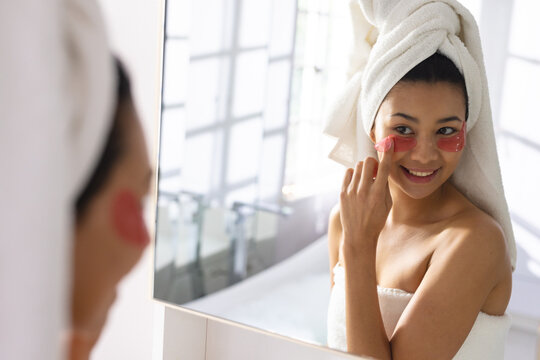 Image of biracial woman with towel on head applying eye mask in bathroom