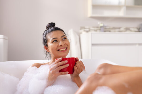 Image of biracial woman relaxing with cup of tea in bathtub in bubble bath in bathroom - Powered by Adobe