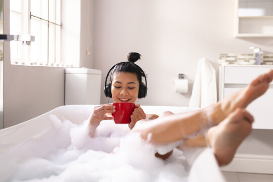 Image of biracial woman relaxing with cup of tea and headphones in bathtub in bubble bath