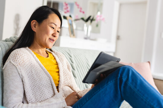 Happy asian woman sitting on sofa in living room and using tablet