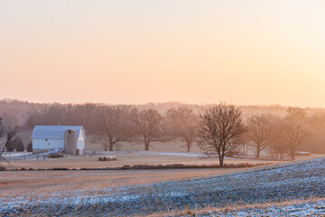 Farm fields with snow on a misty morning in the winter and a small white barn with a silo and round bales. © Margaret Burlingham