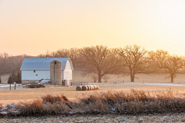 A small, white barn with a silo on a foggy morning in the winter and round bales of hay in a field.