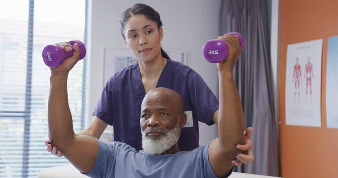 Diverse female physiotherapist and senior male patient holding dumbbells at physical therapy session