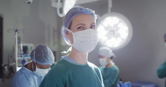 Video portrait of caucasian female surgeon in face mask smiling in operating theatre, copy space