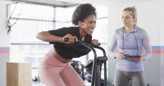 Video of diverse female fitness trainer and woman on exercise bike working out at a gym