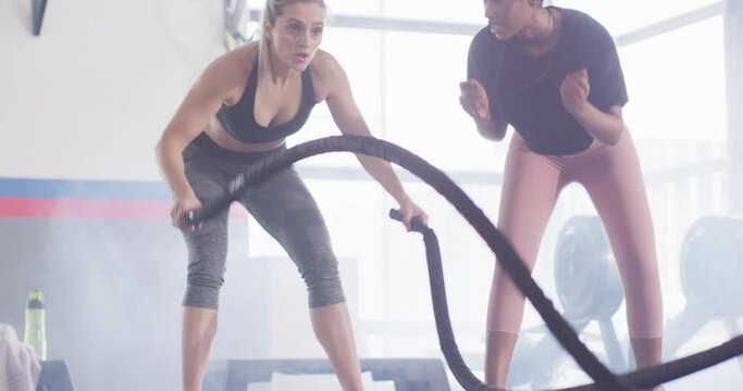 Video of diverse female fitness trainer encouraging woman battling ropes working out at a gym
