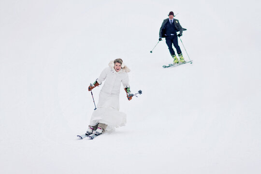 A newlywed couple ski together down a mountain after getting married.
