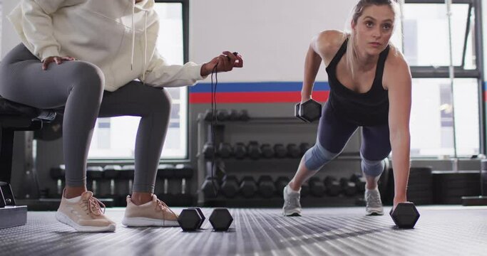 Video of diverse female fitness trainer timing determined woman raising dumbbells working out at gym