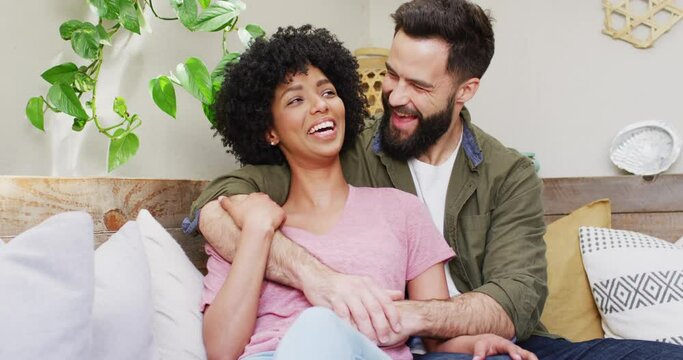 Video portrait of happy diverse couple sitting in living room embracing and smiling